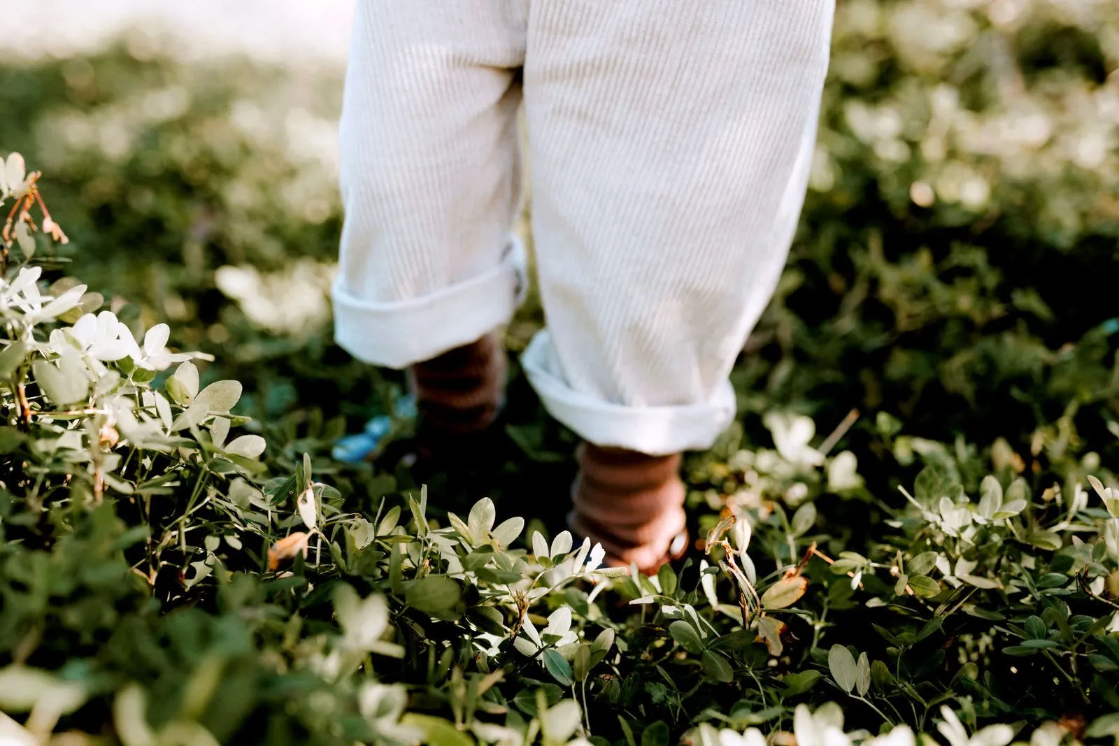 Unrecognizable little kid in white pants standing on green field with foliage in nature against blurred background on sunny weather